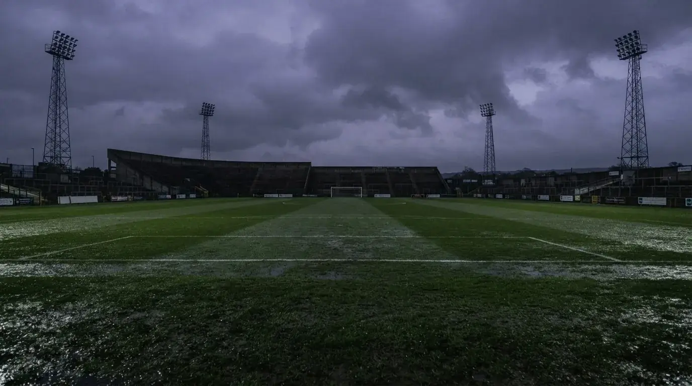 Ein Fußballplatz mit Flutlichtmasten in der Abenddämmerung, aus der Ferne gesehen, leere Tribünen und nasser Rasen unter bewölktem Himmel.