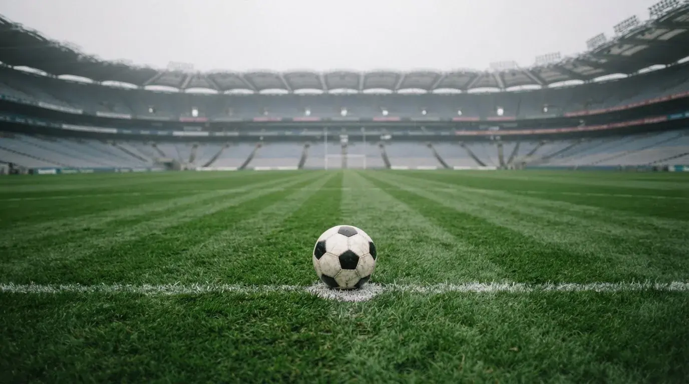 Ein Fußball liegt auf dem Elfmeterpunkt eines leeren Rasenplatzes, im Hintergrund eine Tribüne im Dunst.