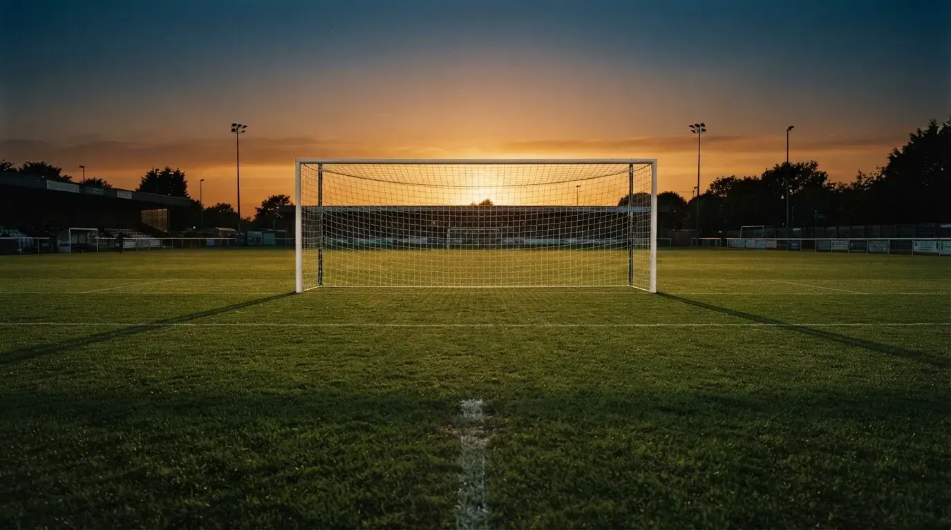 Ein Fußballtor mit Netz bei Sonnenuntergang auf einem leeren Platz, die Schatten der Torpfosten liegen lang auf dem Gras.