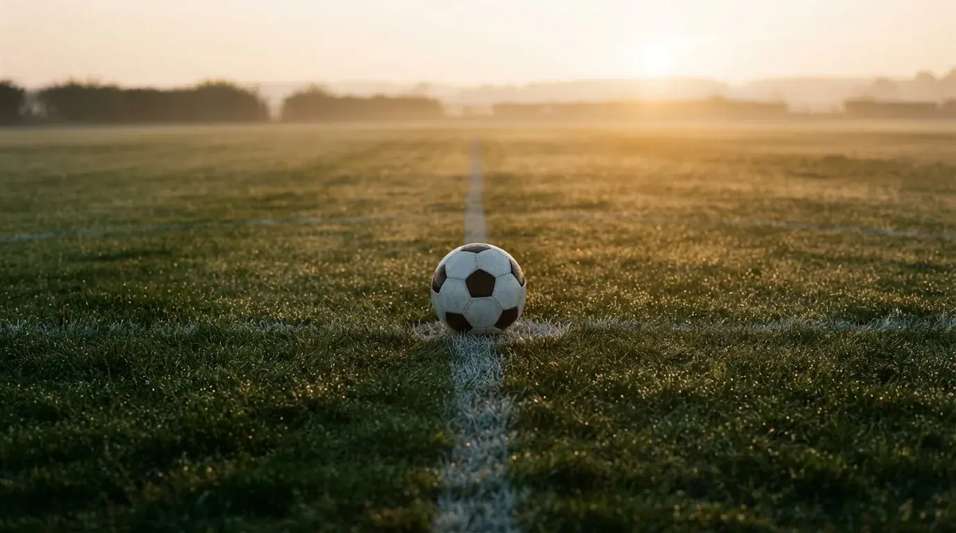 Ein Fußball liegt auf dem Anstoßpunkt eines Rasens bei Sonnenaufgang, Tau glitzert auf den Halmen.