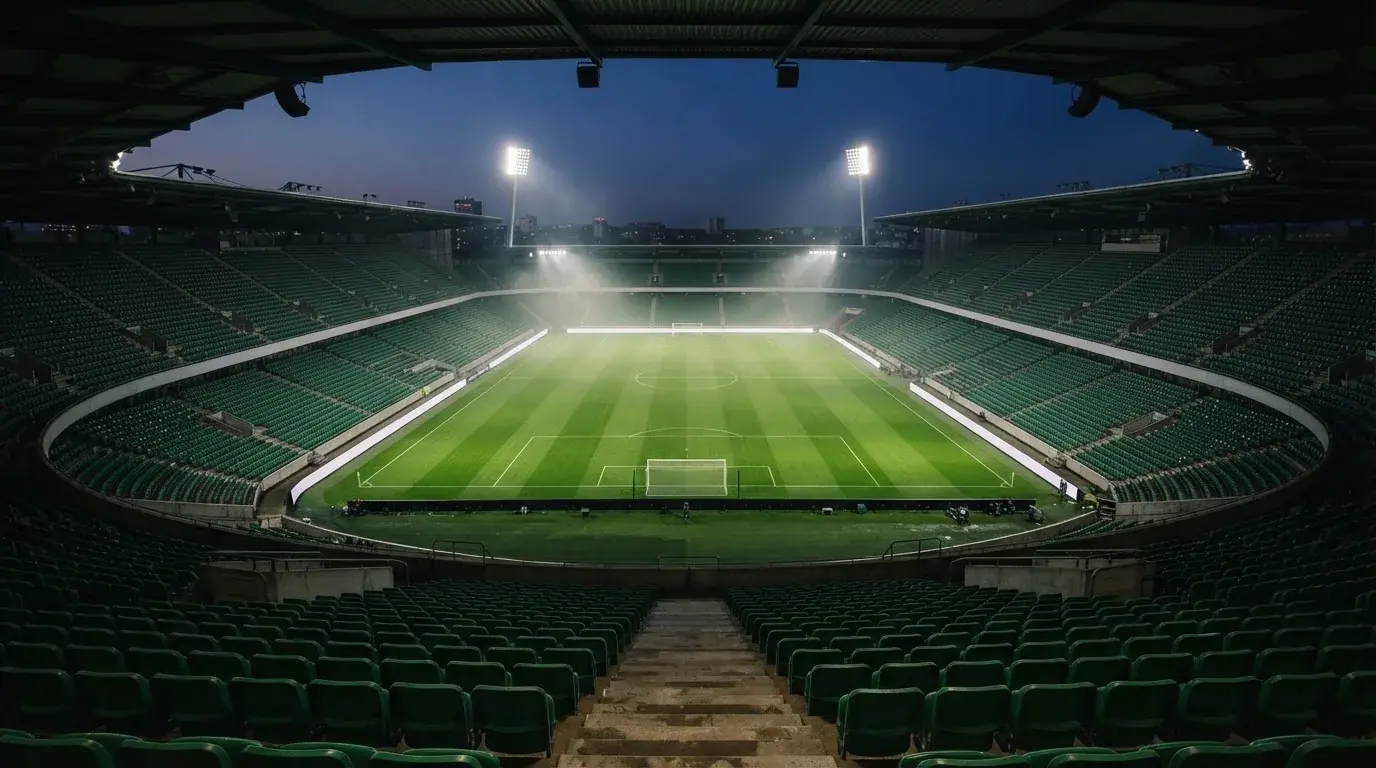 Eine leere Stadiontribüne unter Flutlicht aus der Ferne betrachtet, die Rasenfläche leuchtet grün in der Dämmerung.