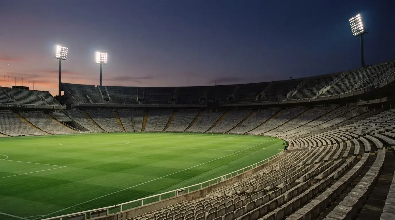 Eine leere Stadiontribüne unter Flutlicht aus der Ferne betrachtet, die Rasenfläche leuchtet grün in der Dämmerung.