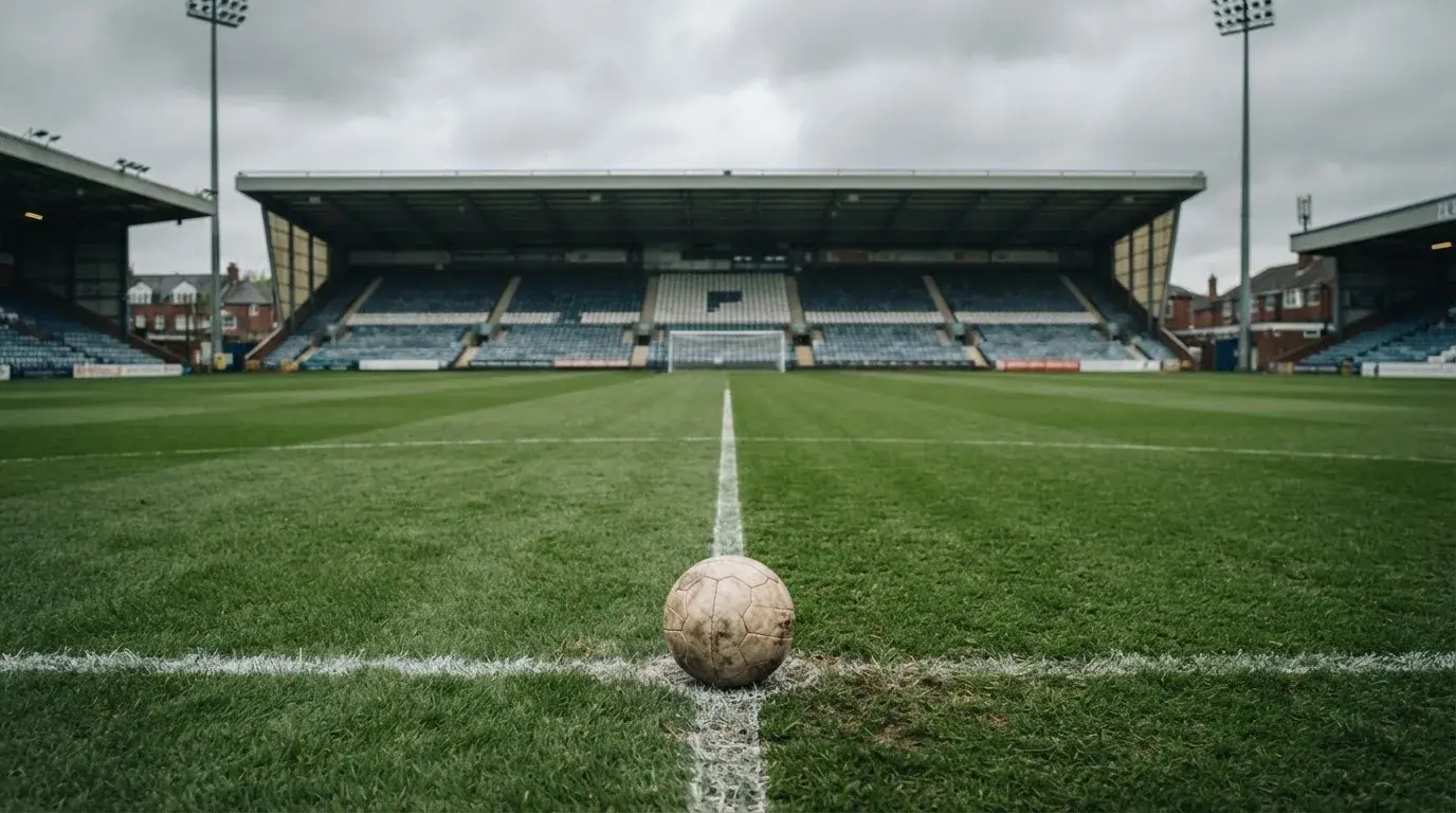 Ein Fußball liegt auf dem Anstoßpunkt eines leeren Rasenplatzes, im Hintergrund eine ruhige Tribüne unter bedecktem Himmel.