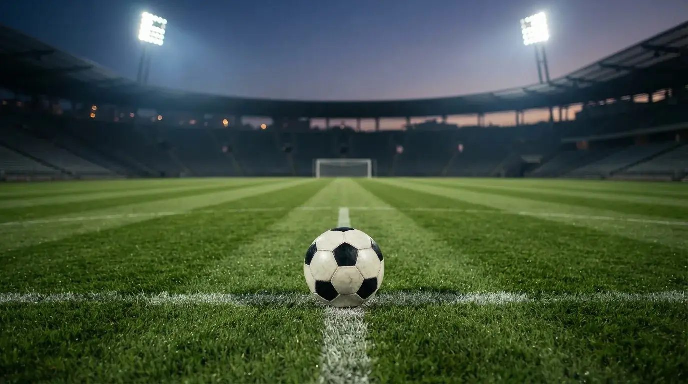 Ein Fußball liegt auf dem Rasen eines leeren Platzes, im Hintergrund ein Stadion unter Flutlicht in der Abenddämmerung.