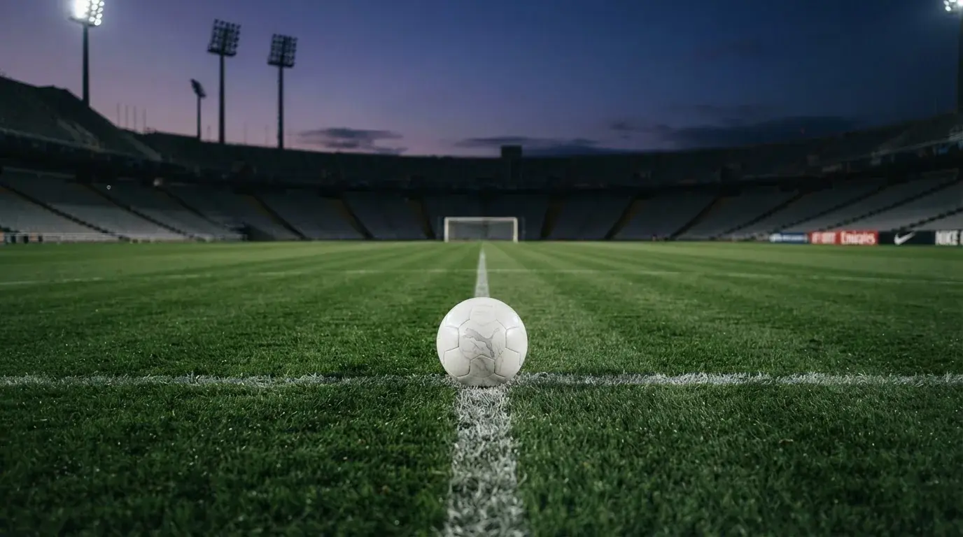 Ein Fußball liegt auf dem Anstoßpunkt eines leeren Rasenplatzes, im Hintergrund steht ein Stadion unter Flutlicht in der Dämmerung.