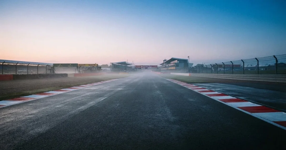 Eine leere Rennstrecke mit rot-weißen Randsteinen im frühen Morgenlicht, auf dem Asphalt liegt dünner Nebel, keine Fahrzeuge.